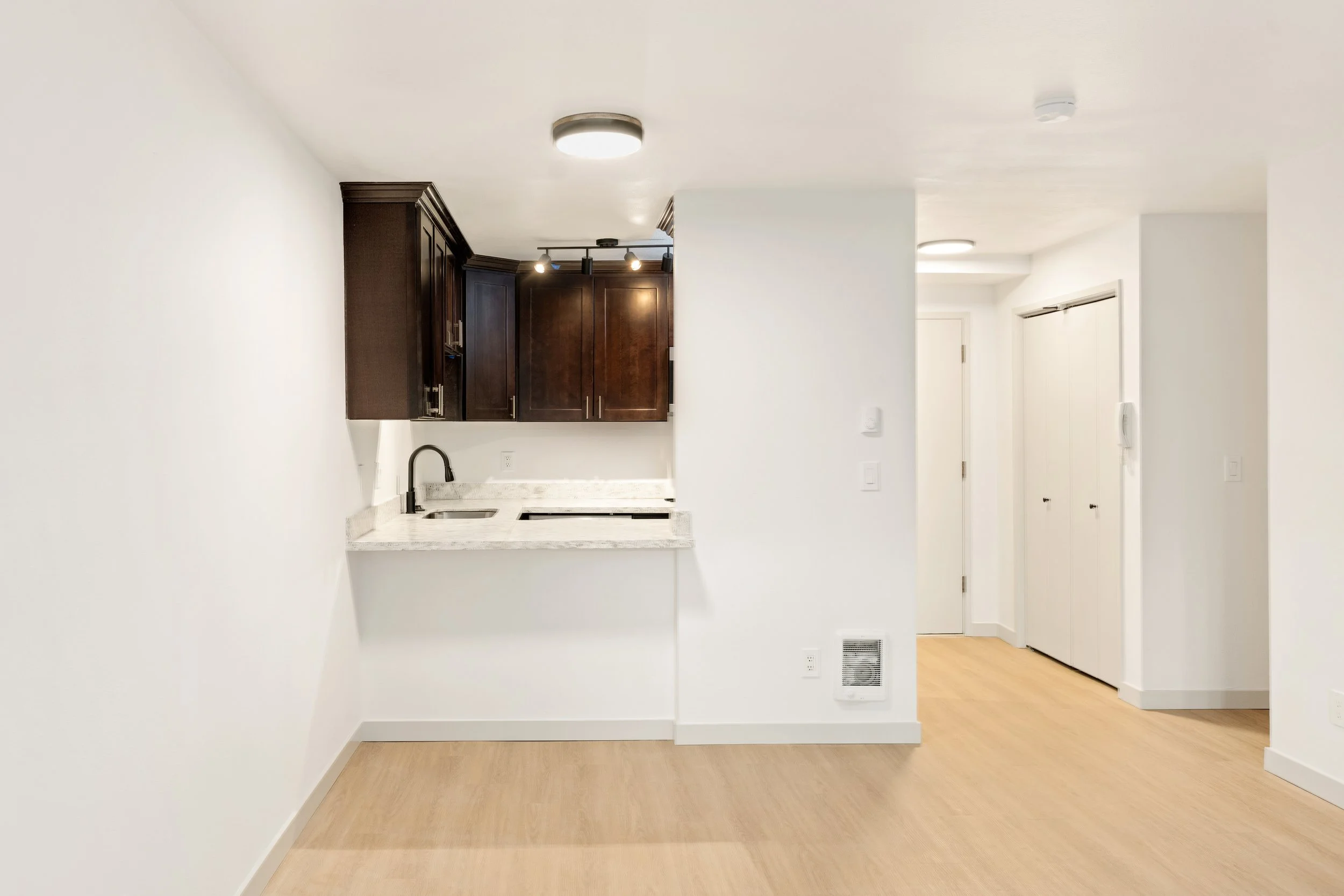 Empty living area with light wood flooring, white walls, and a small kitchen with dark brown cabinets and a white granite countertop.
