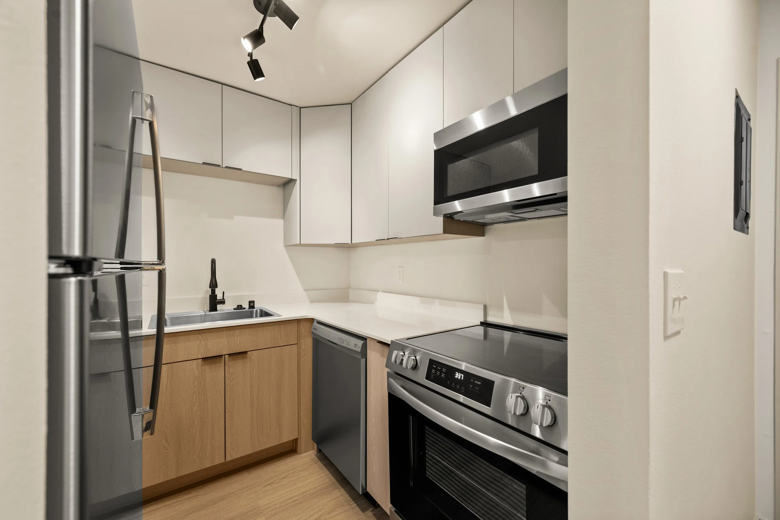 Modern kitchen with stainless steel refrigerator, black microwave, electric stove, dishwasher, white countertops, and wooden cabinets.