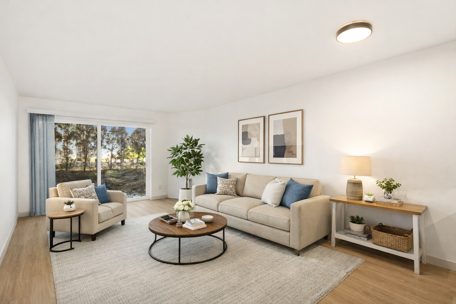 Bright and airy living room with beige sofa, matching armchair, wooden coffee table, side table, and a white console table with plants, lamp, and art on the wall, large window with curtains showing trees outside.