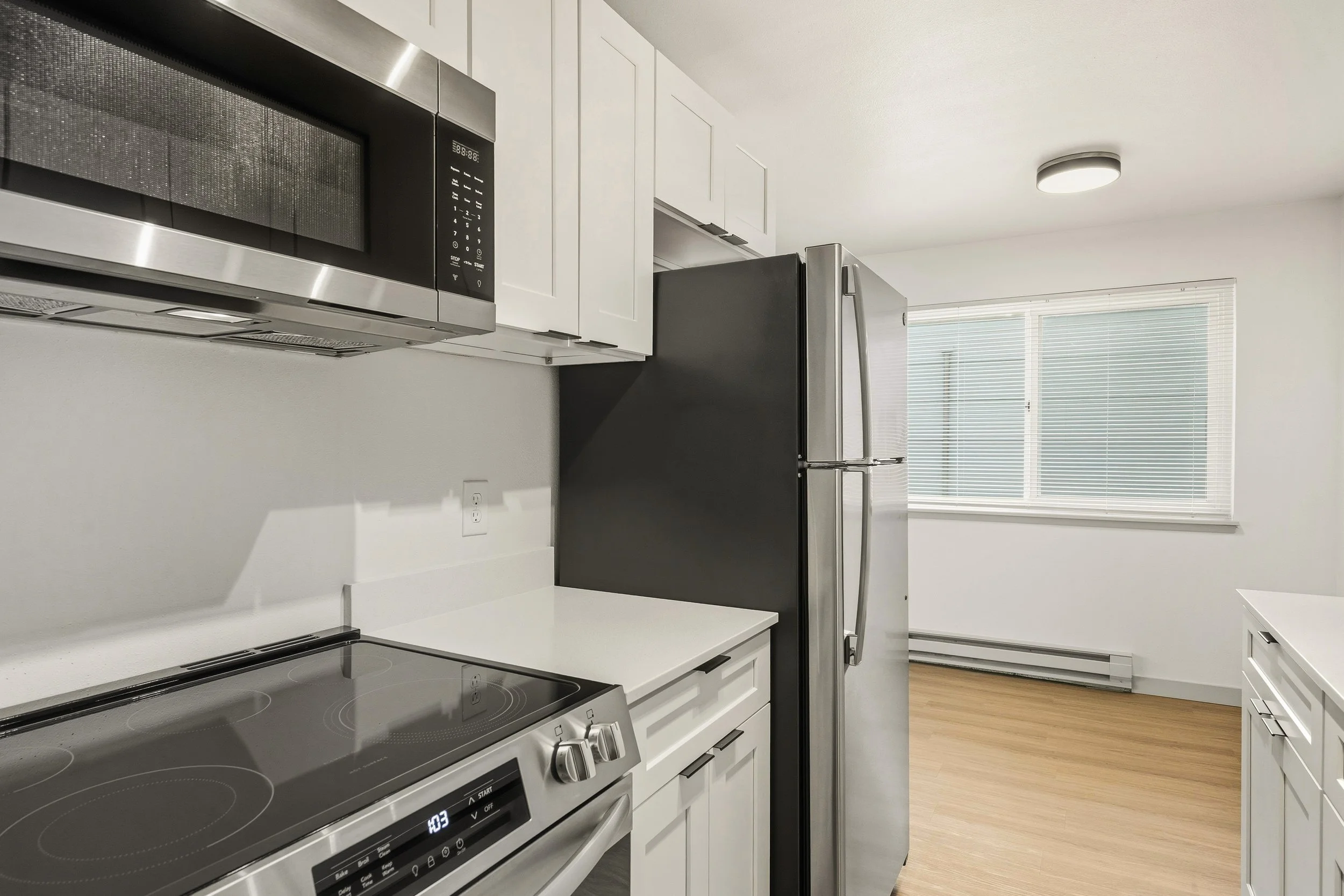 Kitchen with white cabinets, stainless steel microwave, black refrigerator, electric stove, and large window with blinds.