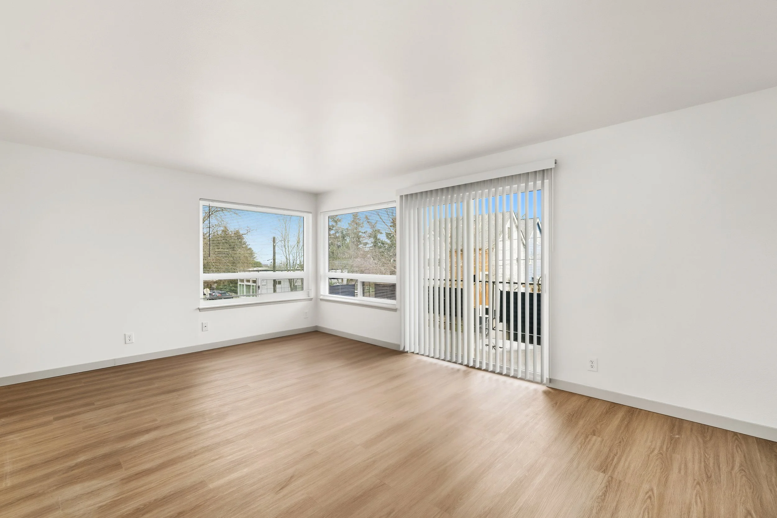 Empty living room with white walls, hardwood floors, large windows with blinds, and sliding glass doors with vertical blinds leading to an outdoor area.