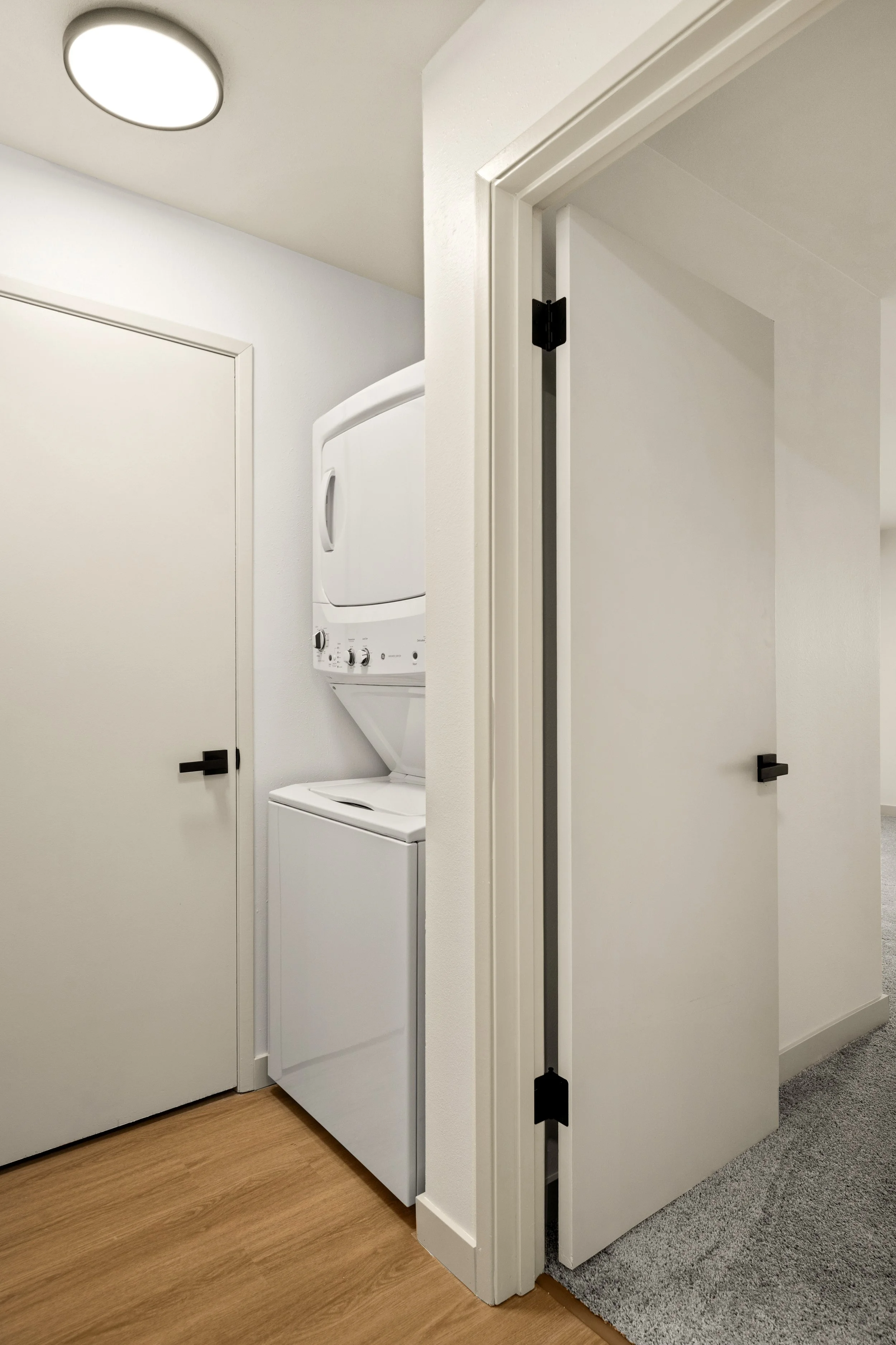 A laundry area with a stacked washer and dryer next to a closed door, with another partially open white door on the right side, in a room with a wood and carpeted floor.