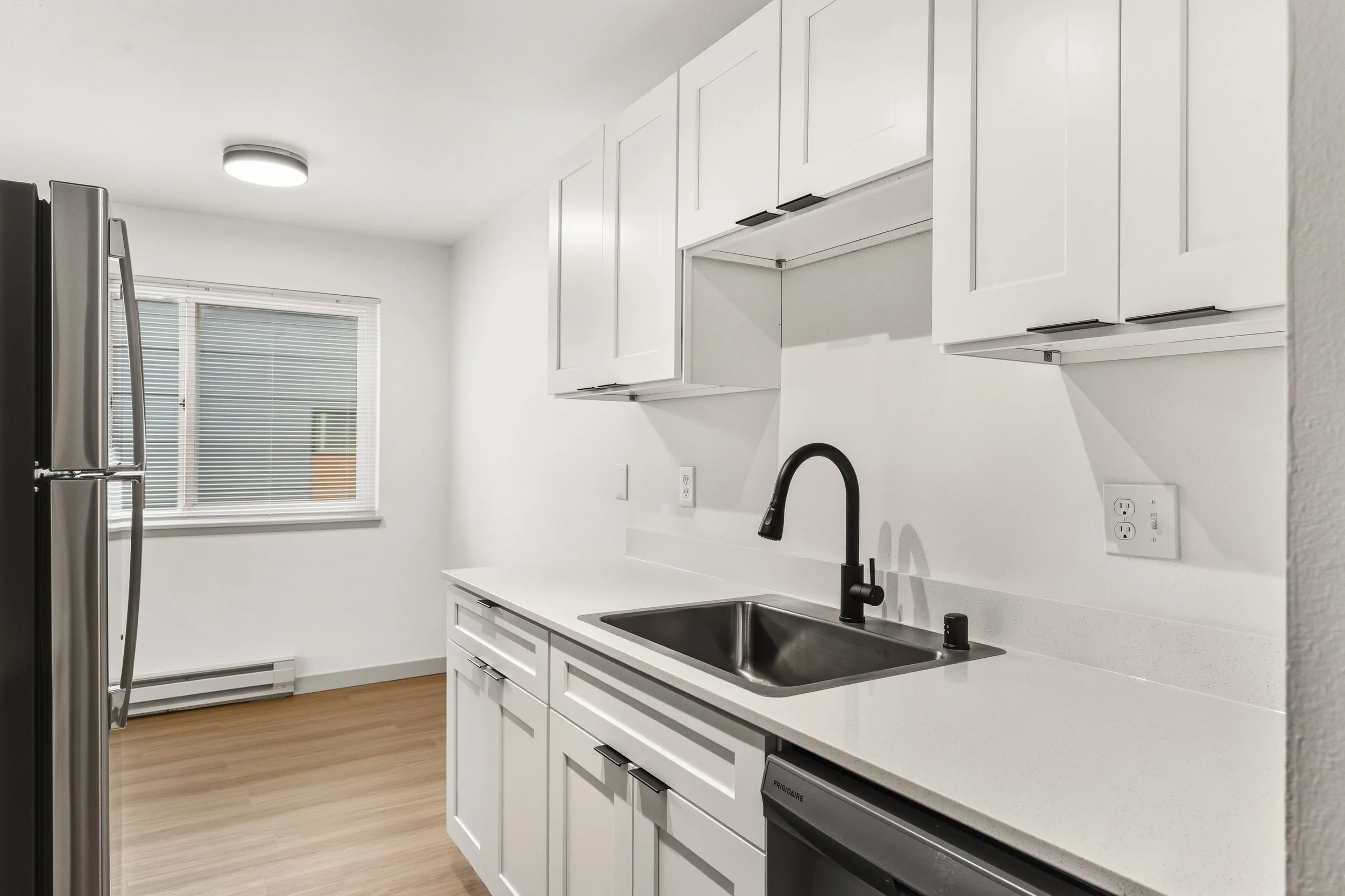 Modern kitchen with white cabinets, a stainless steel refrigerator, a black faucet, and light wood floors, illuminated by a ceiling light