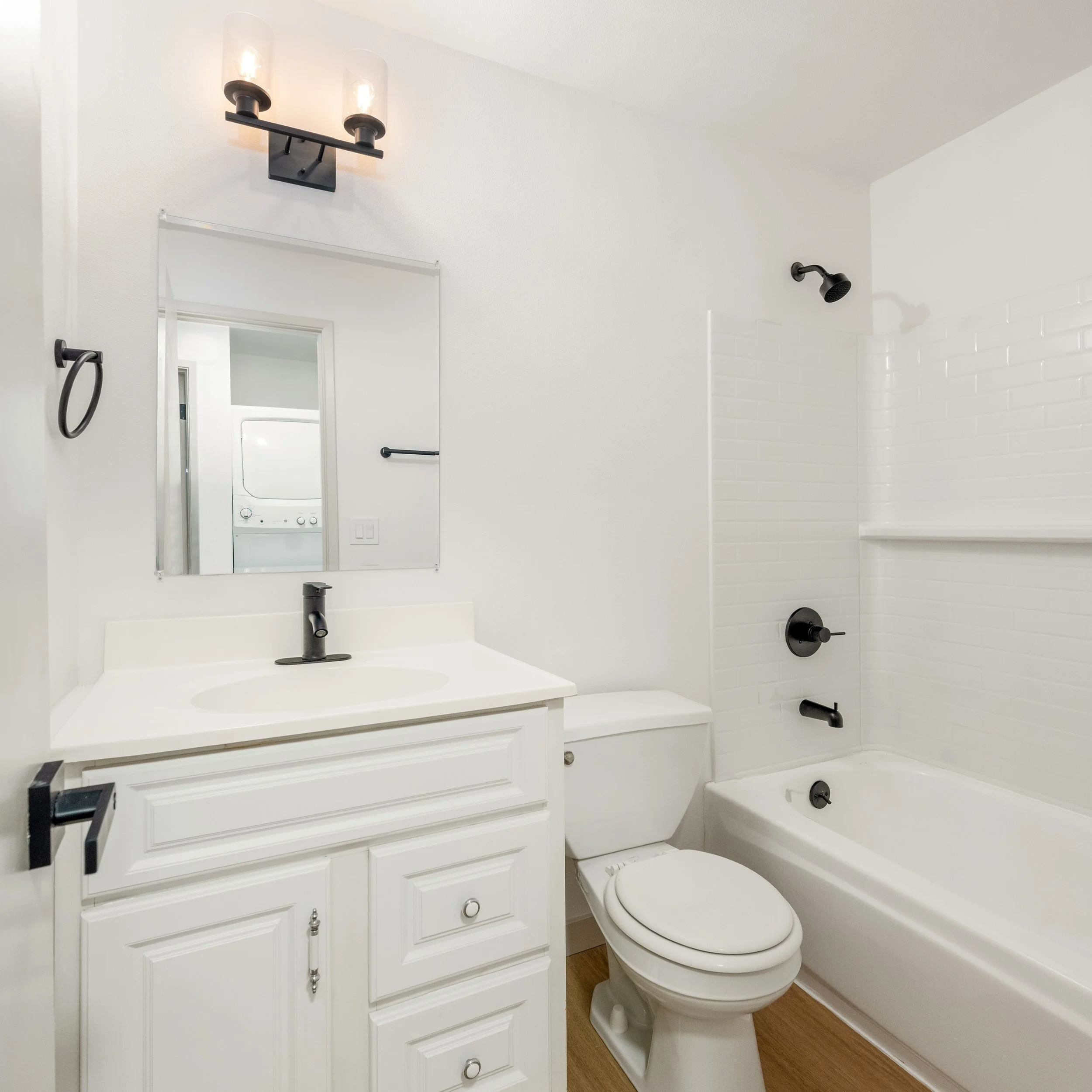 A small bathroom with a white vanity and mirror, a toilet, and a bathtub with a shower. black fixtures, white walls, and a light wood floor.