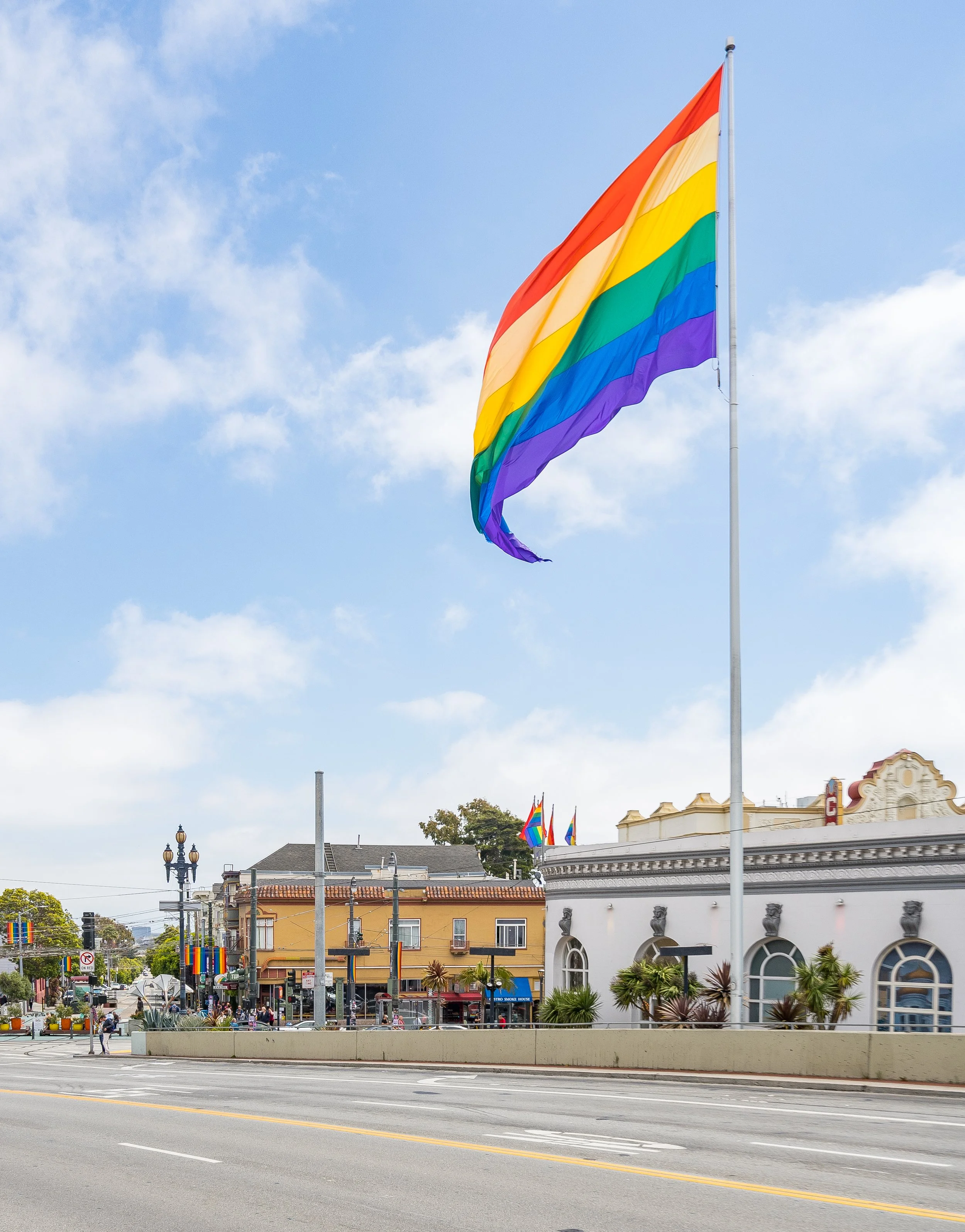 Large rainbow pride flag flying on a flagpole in a city street with smaller rainbow flags on buildings, and a white building with decorative architecture, blue sky with clouds in the background.