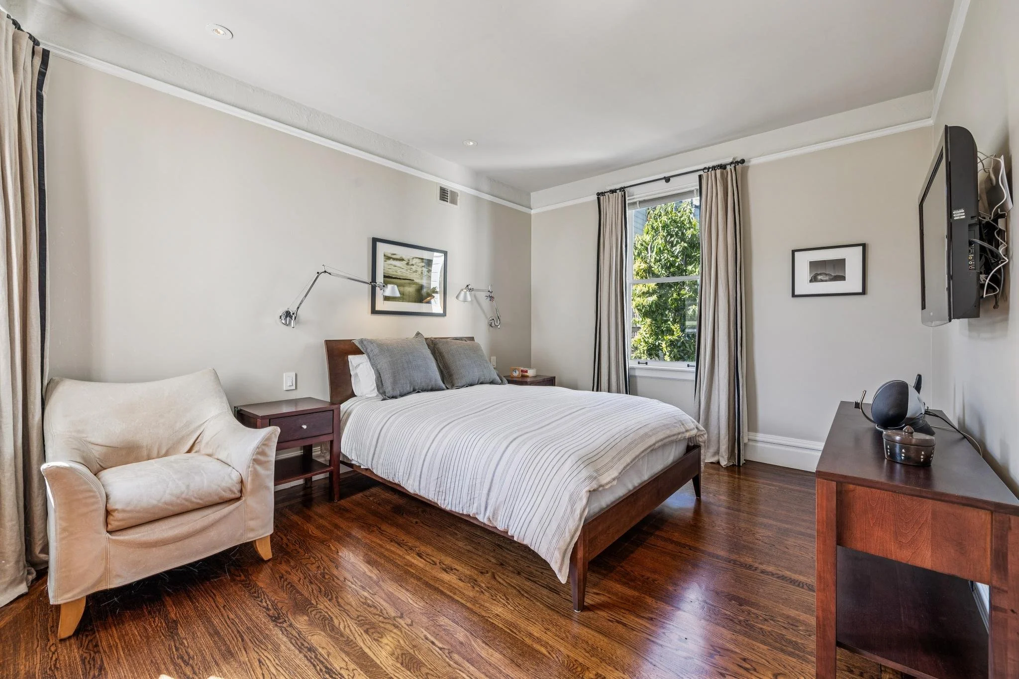 A bedroom with a bed, a cream armchair, a nightstand, a dresser with a TV, and a window with curtains