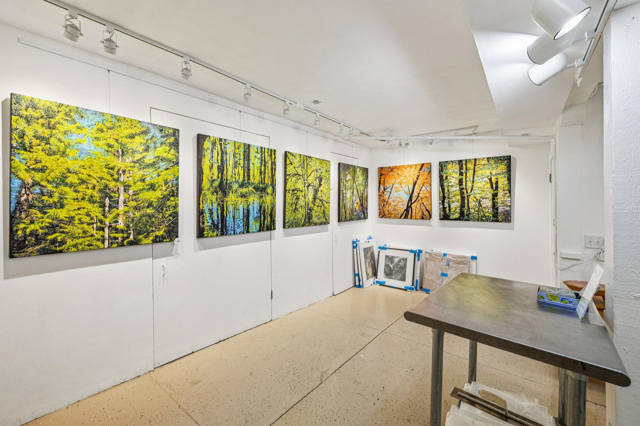 Gallery with landscape paintings of trees and nature, displayed on white walls, with frames leaning against the wall on the floor, and a table with an informational sign in the foreground.