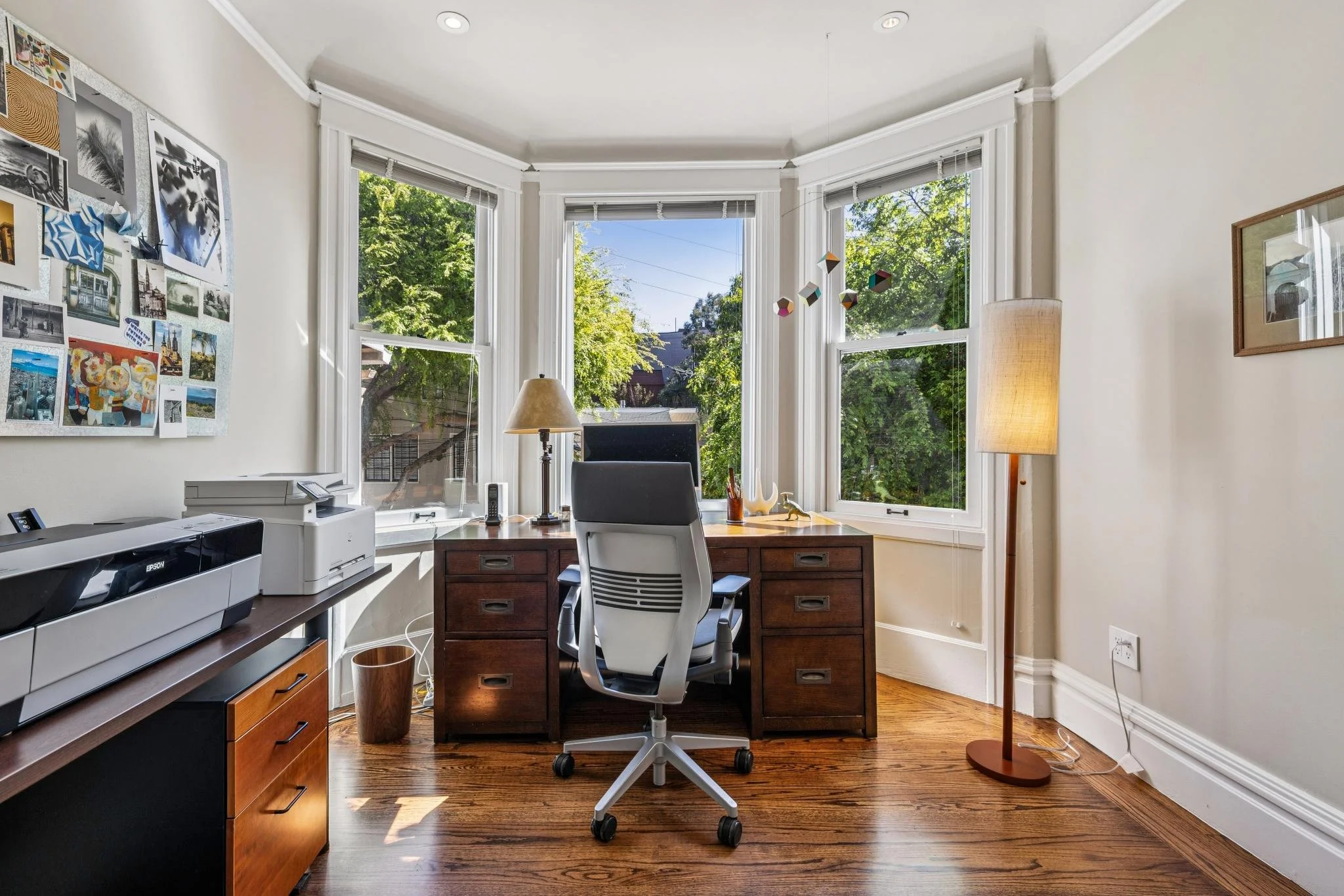 Home office with a wooden desk, white swivel chair, computer monitor, desk lamp, and a window view of green trees outside.