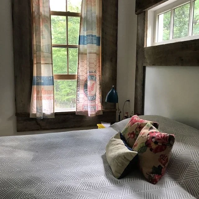 A cozy bedroom with a large window covered by curtains, a bed with white bedding and two floral pillows, and a small blue bedside lamp.