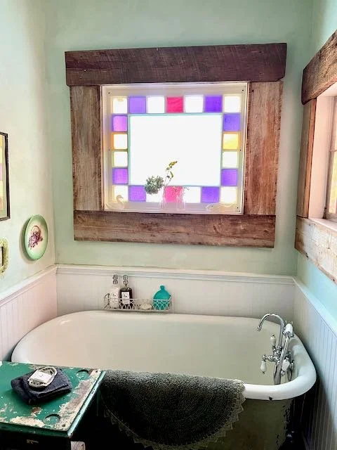 A vintage-style bathroom with a white clawfoot tub placed under a decorative stained glass window with purple, pink, and yellow panes and a small potted plant on the window sill. A side table with a telephone and a gray towel hangs over the tub. The 