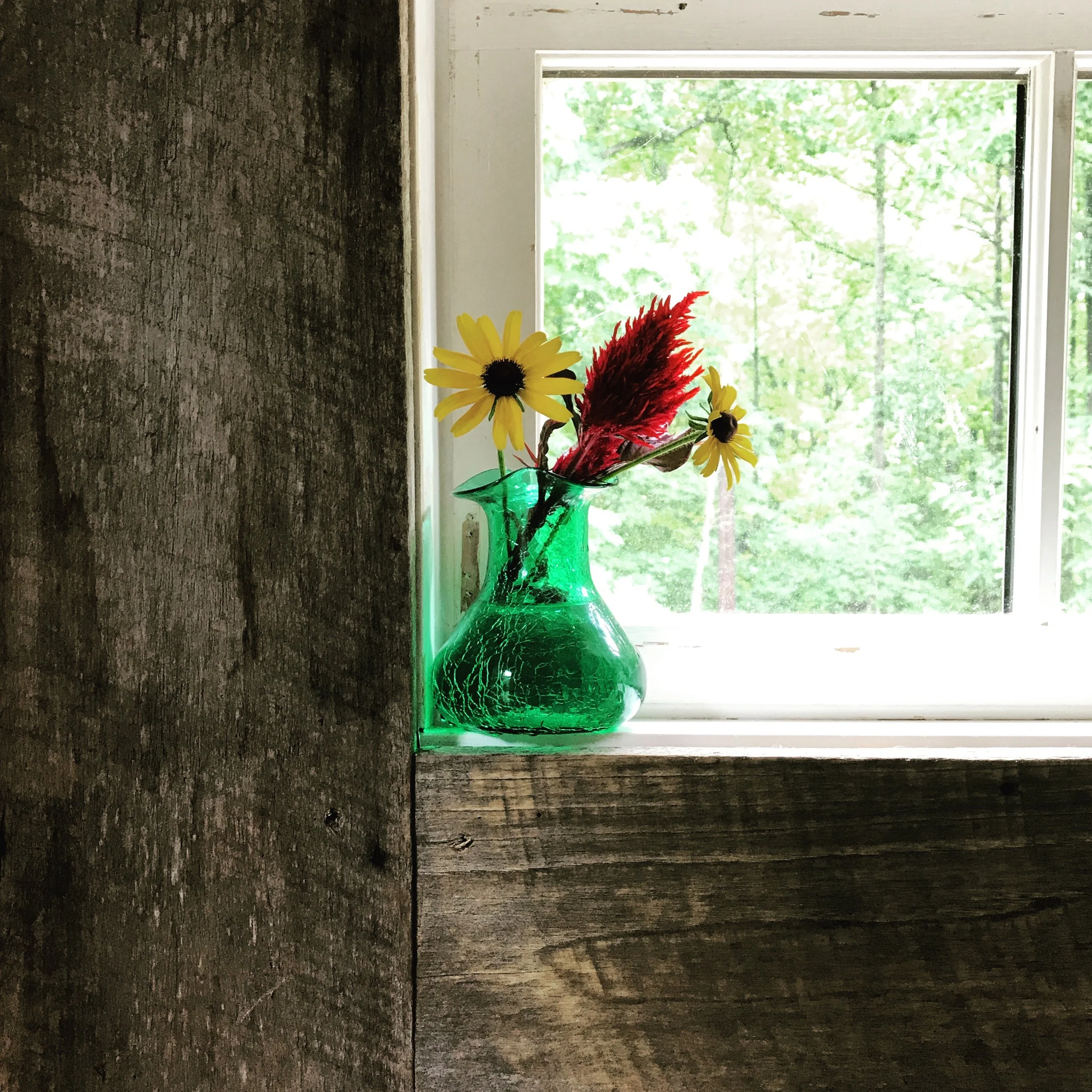 A green glass vase on a window sill holding yellow daisies and red dried grass with a background of a wooded landscape visible through the window.