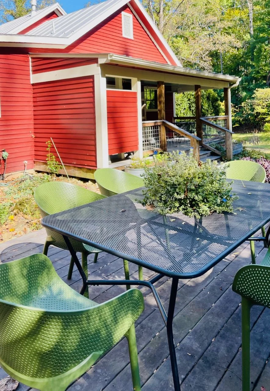 Outdoor patio with green chairs around a black metal table and a potted plant on top, in front of a red house with a covered porch and a wooden staircase, surrounded by trees and greenery.