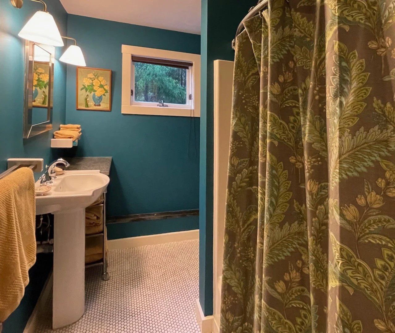 A small bathroom with teal walls, a white pedestal sink, a mirror with lamps, a window showing greenery outside, a framed floral painting, a towel rack with a yellow towel, and a shower with a patterned curtain featuring green leaves.