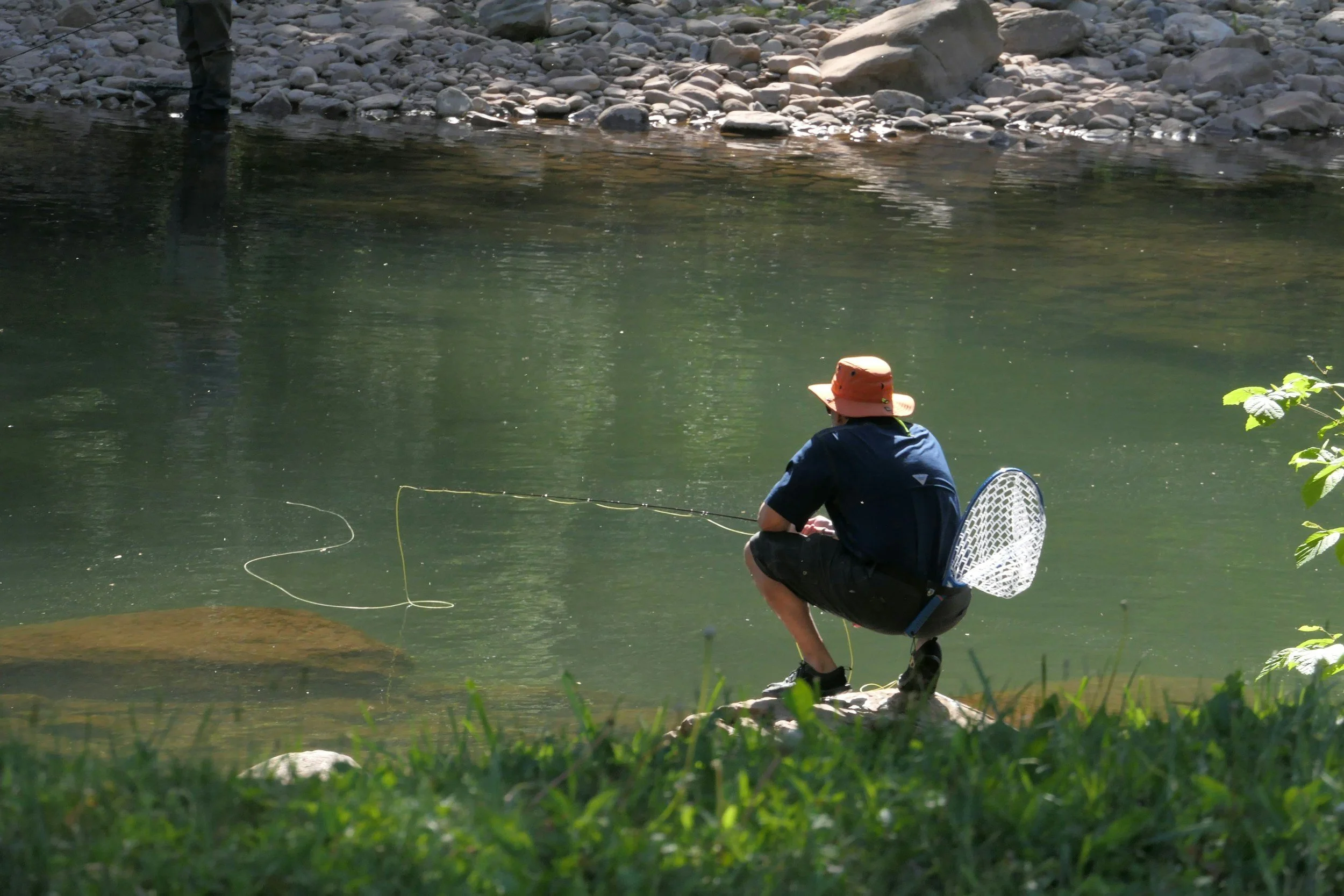 A person in a dark shirt, shorts, and orange hat crouches on a rock at the edge of a river, fishing with a fishing rod and a net attached to their back. The river has rocks along the shoreline and the water is calm.