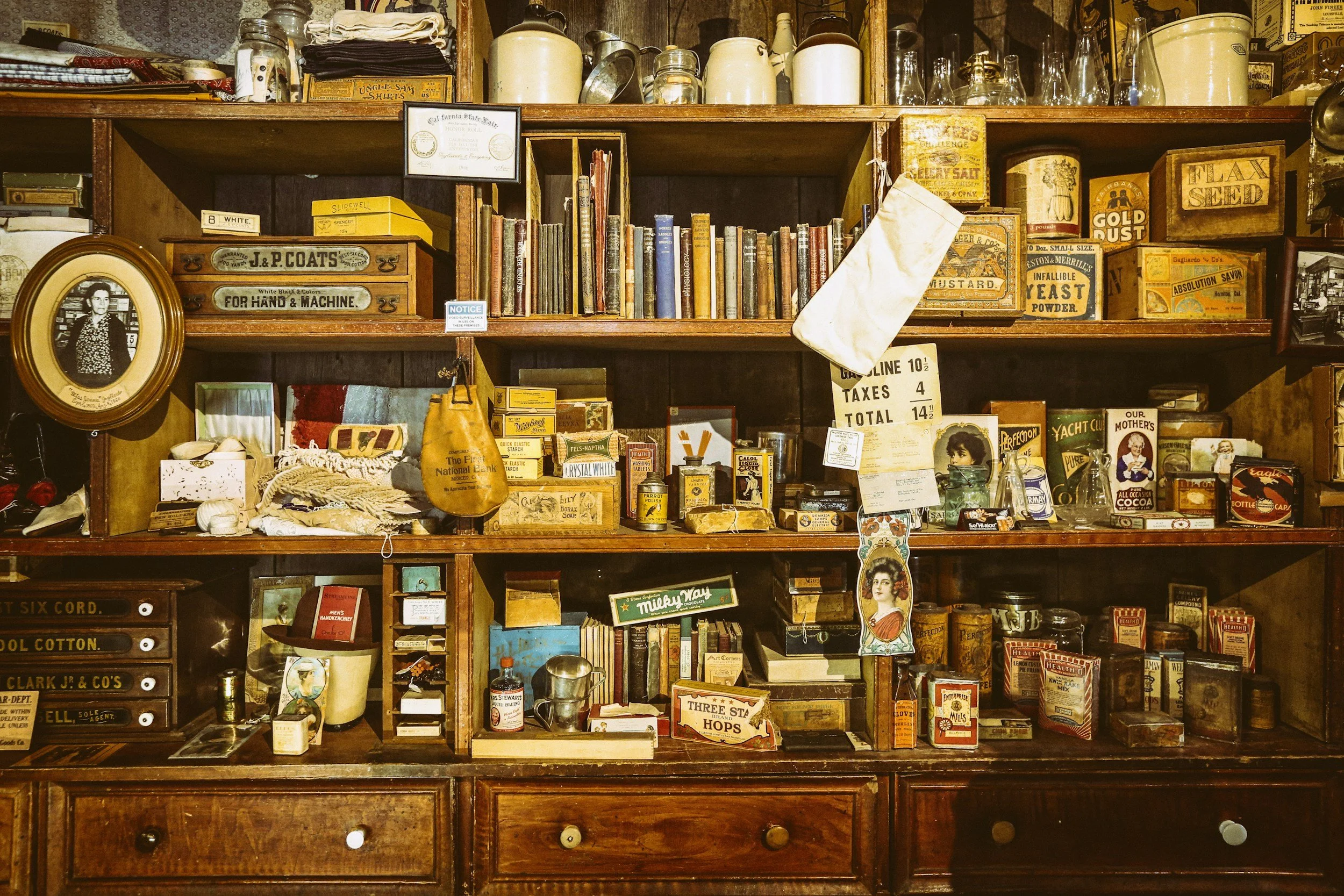A wooden wall unit filled with vintage items, books, old-fashioned tins, glass bottles, and miscellaneous collectibles in an antique store.