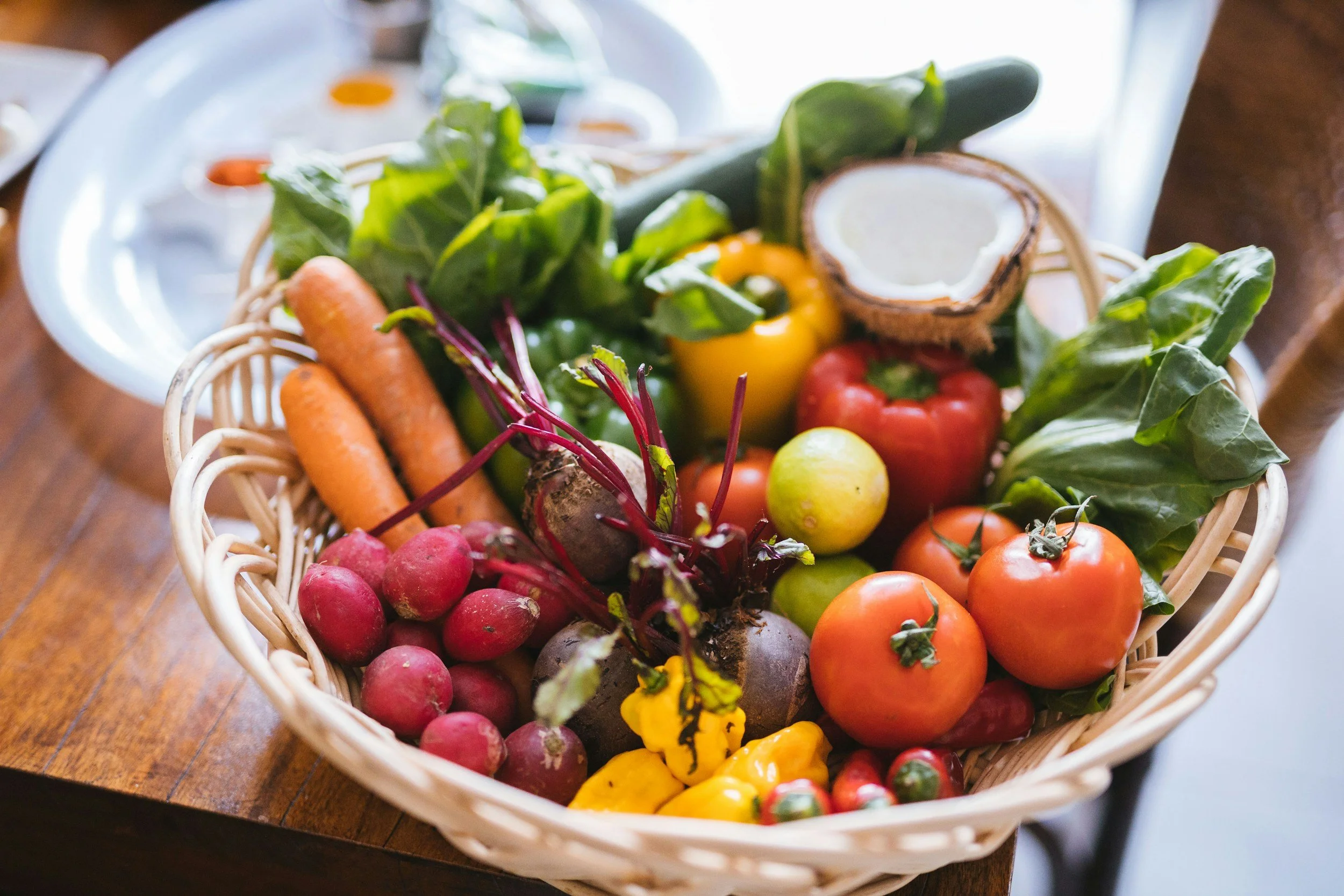 A basket filled with fresh vegetables including carrots, radishes, beets, tomatoes, bell peppers, lettuce, cucumber, and a halved coconut with white flesh, placed on a wooden table.