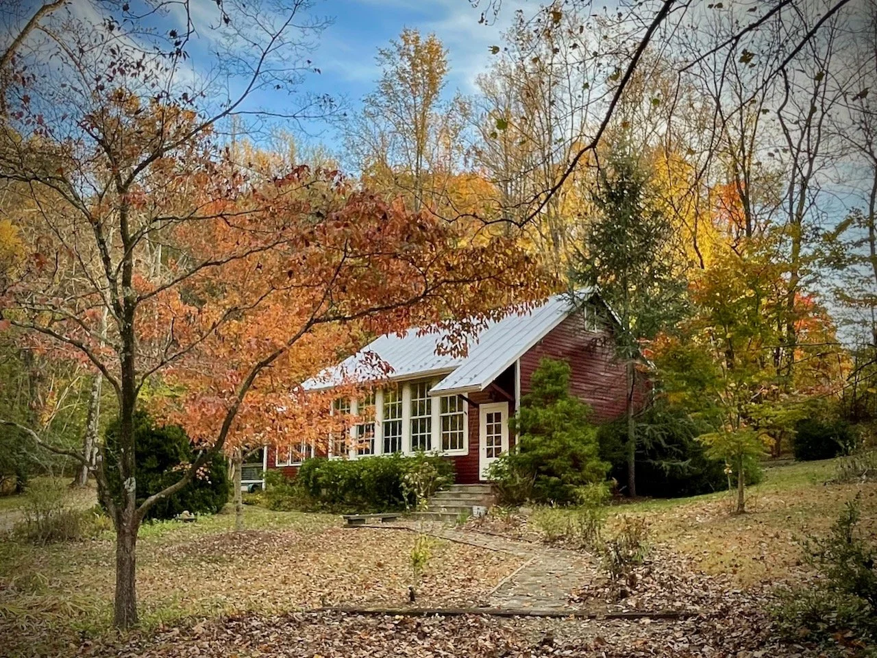 Red house with white trim and large windows surrounded by colorful autumn trees, fallen leaves on the ground, and a cloudy sky.