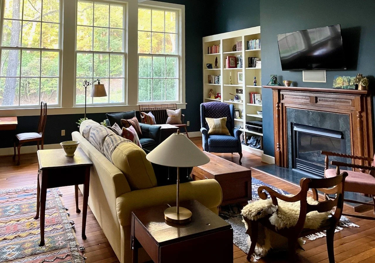 Living room with large window, yellow sofa, blue armchair, wooden chairs, built-in bookshelf, fireplace with TV mounted above, and hardwood floors.