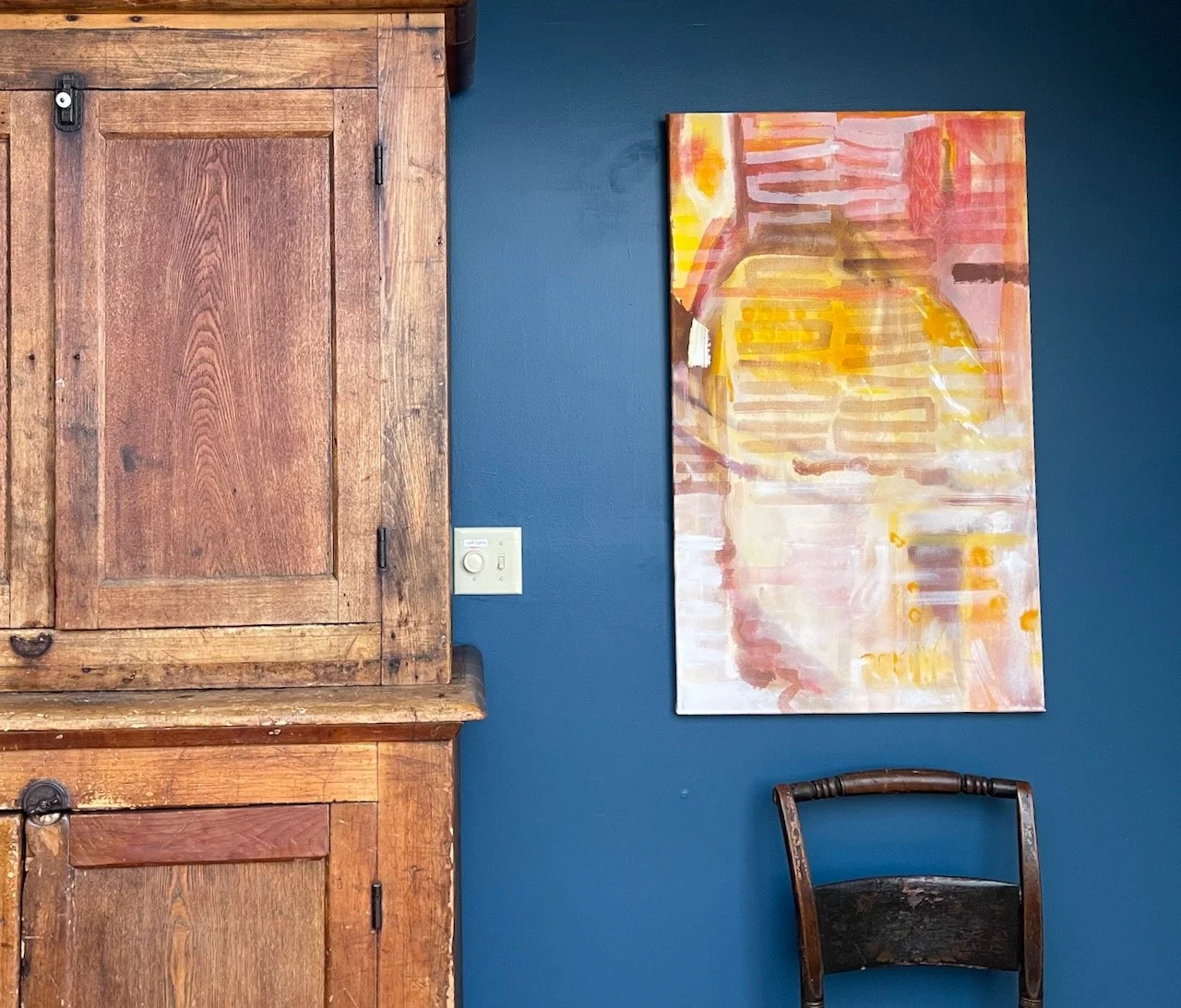A room with a dark blue wall, a wooden cabinet on the left, an abstract painting with warm colors hanging on the wall, and a wooden chair with a dark seat in front of the wall.