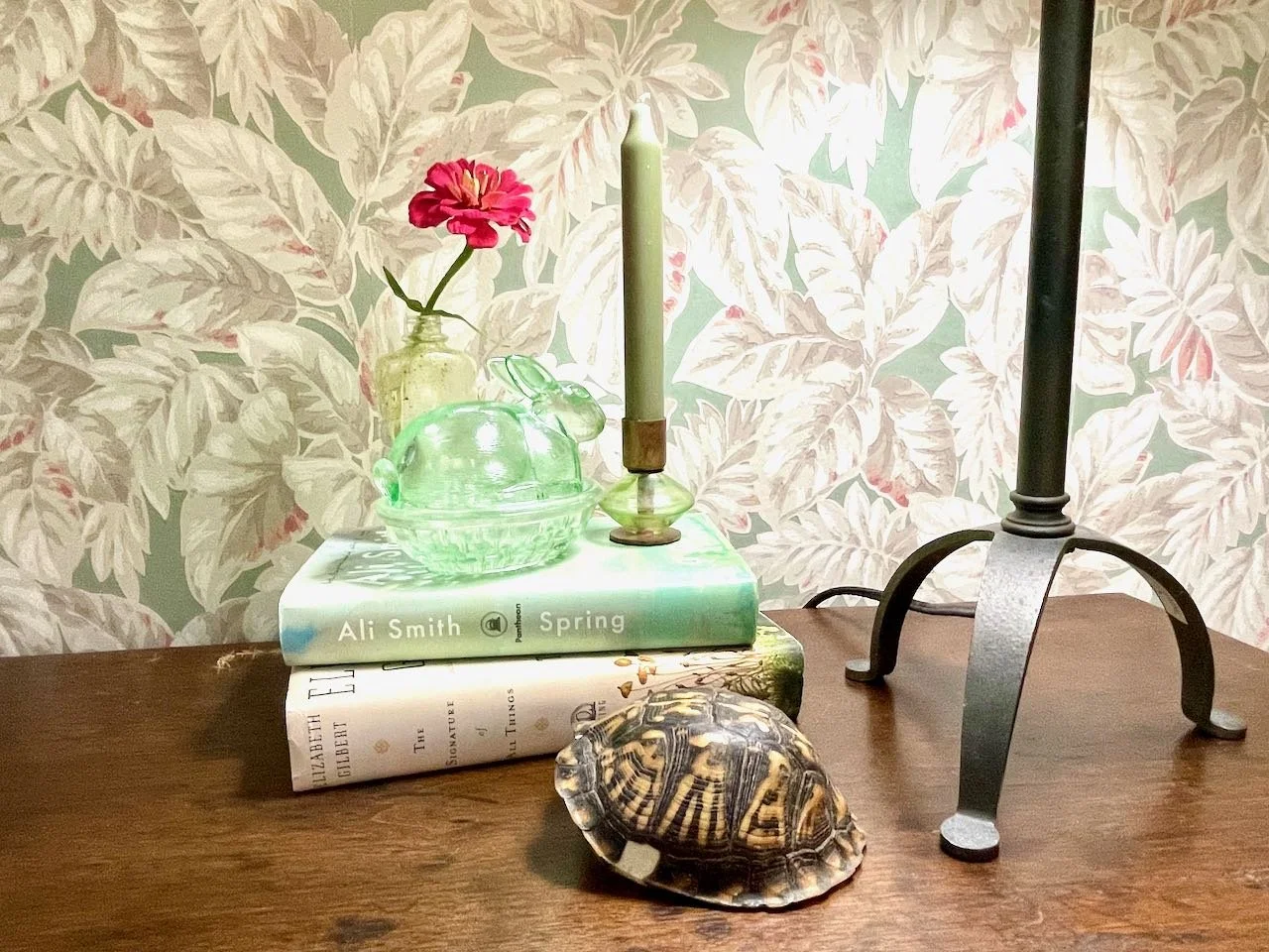Decorative tabletop arrangement with a book, shell, candle, glass turtle figurine, and blotter with a floral wallpaper background.