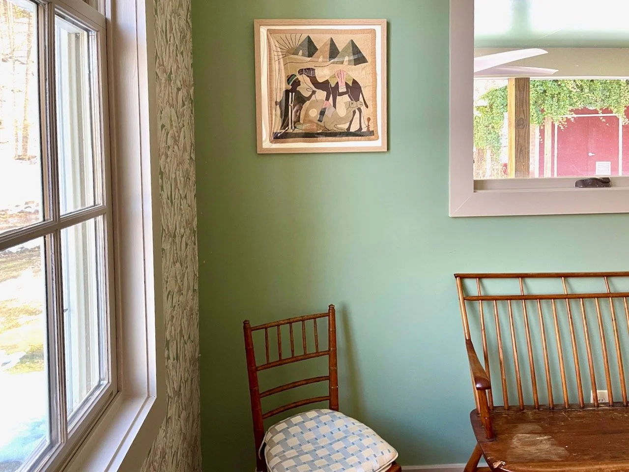 Corner of a room with green wall, wooden chairs, framed artwork, window showing trees and red building outside.