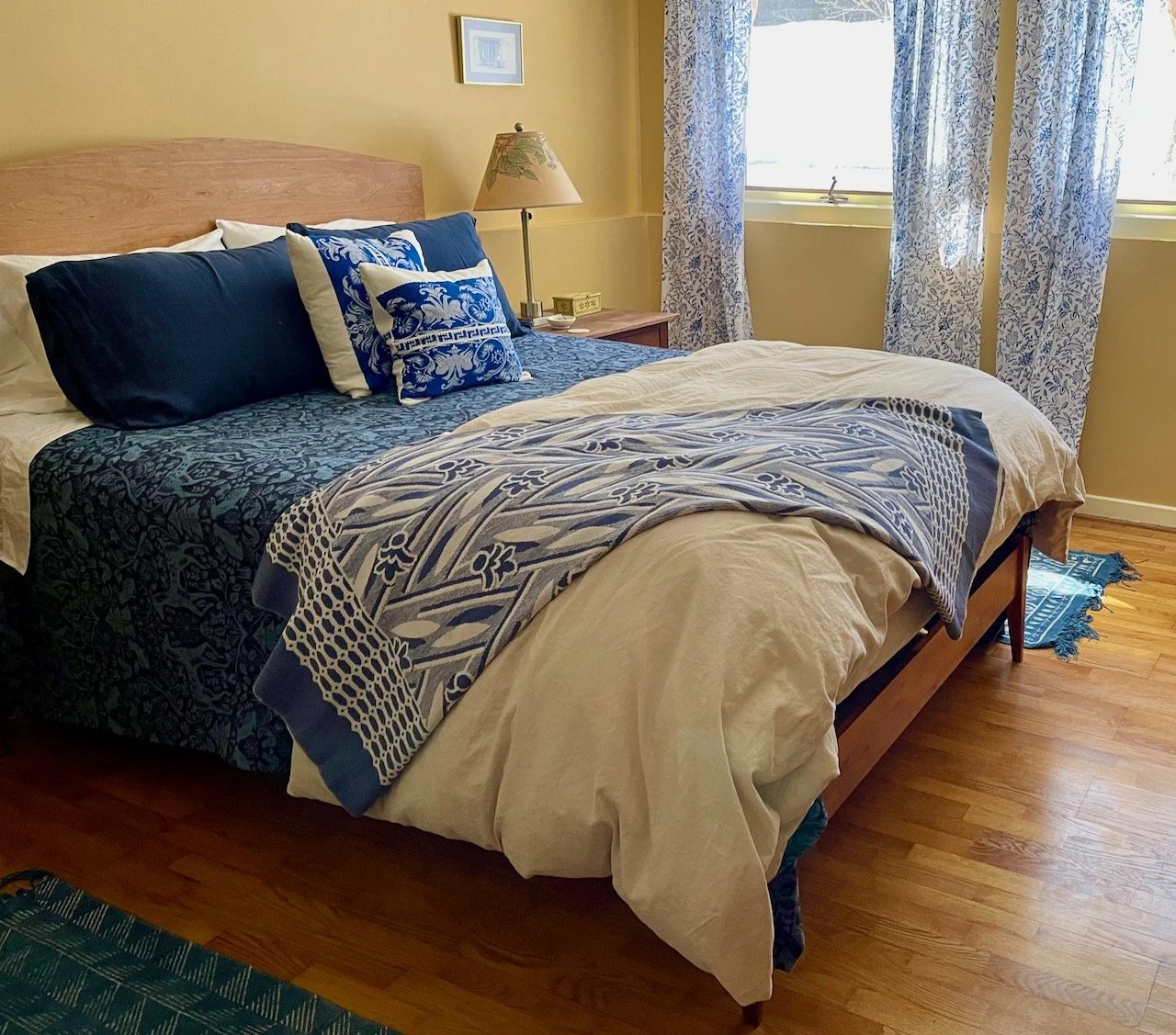 A tidy bedroom with a bed made with navy blue and white bedding, accented with decorative pillows. There is a wooden nightstand with a lamp, a small picture frame on the wall, and windows with patterned curtains letting in natural light. The floor is