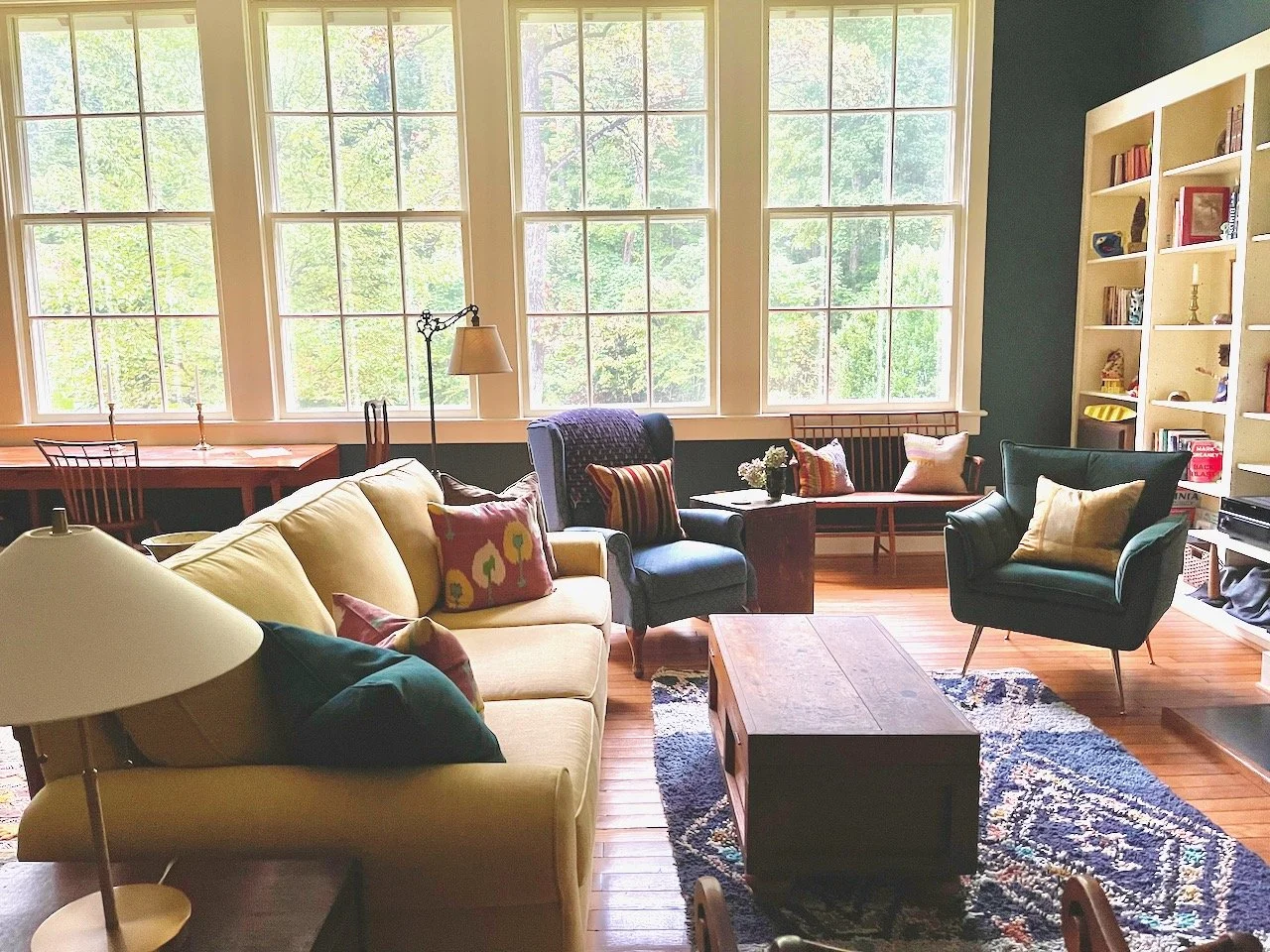 Living room with large windows, yellow sofa, blue armchairs, wooden coffee table, bookshelf, and various throw pillows.