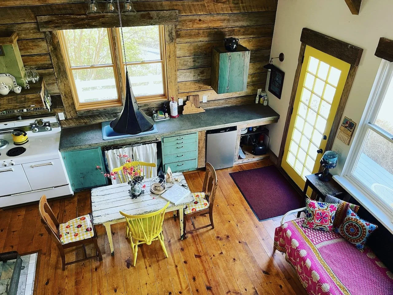 A cozy kitchen and dining area with wooden floors, a yellow door with glass panes, a small white table with three chairs, and a pink patterned sofa with colorful pillows. The kitchen has rustic wood walls, a white oven, a small refrigerator, and gree