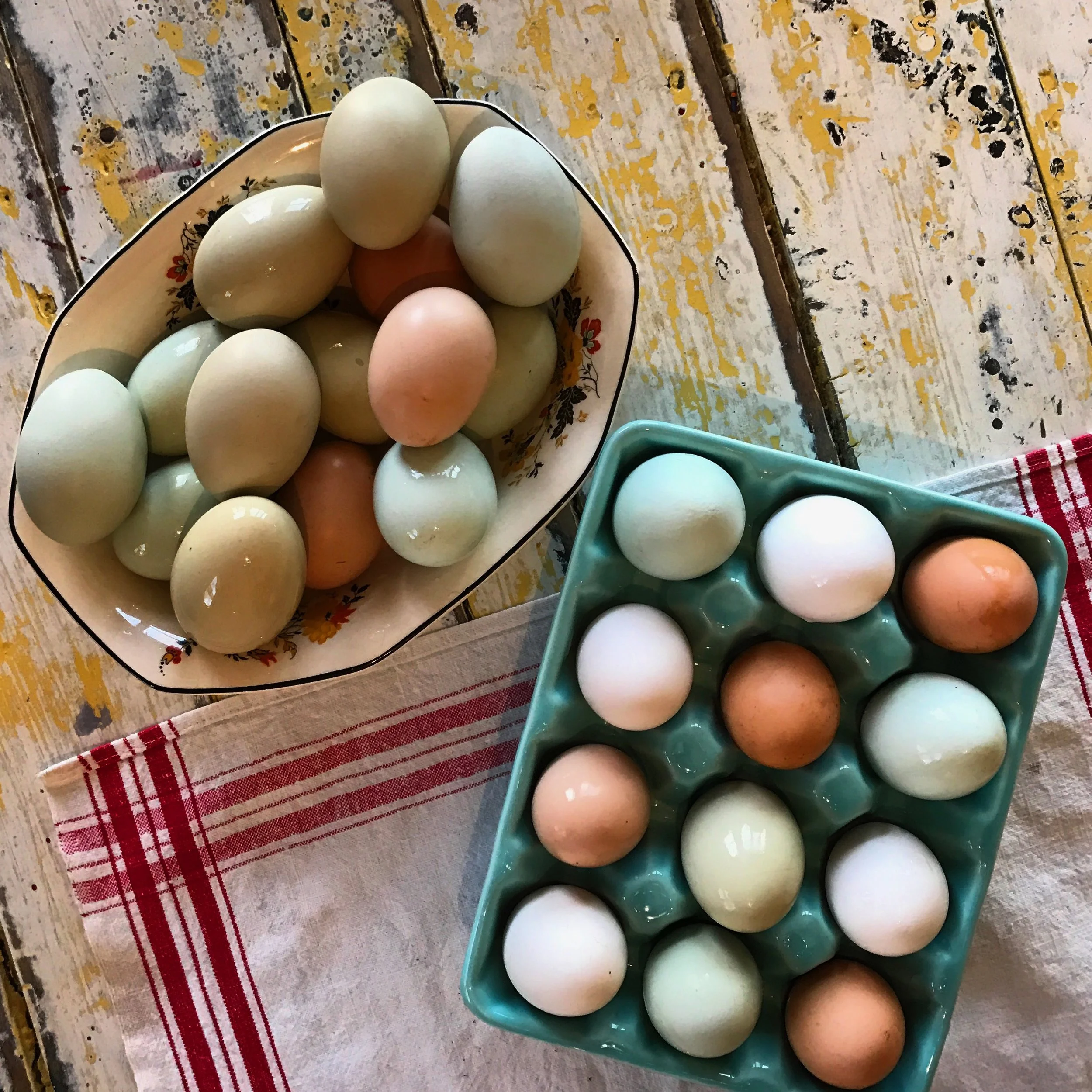 A bowl and a carton of mixed colored eggs placed on a rustic wooden table with a cloth underneath.