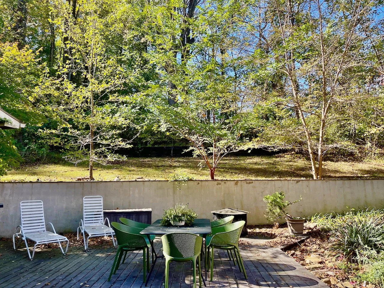 A backyard patio with a round table and six green chairs, two white lounge chairs, a potted shrub, and a view of trees beyond a concrete wall.
