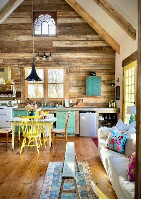 Open-concept rustic kitchen and living area with wood walls and ceiling, yellow dining table, yellow chairs, a black hanging light, a white sofa with colorful pillows, hardwood floors, a small refrigerator, and large windows letting in natural light.