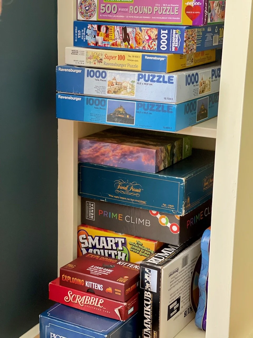 Shelf filled with various board games and puzzles, including 1000-piece Ravensburger puzzles, a 500-piece puzzle, and smaller games like Exploding Kittens, Scrabble, and Prime Climb.