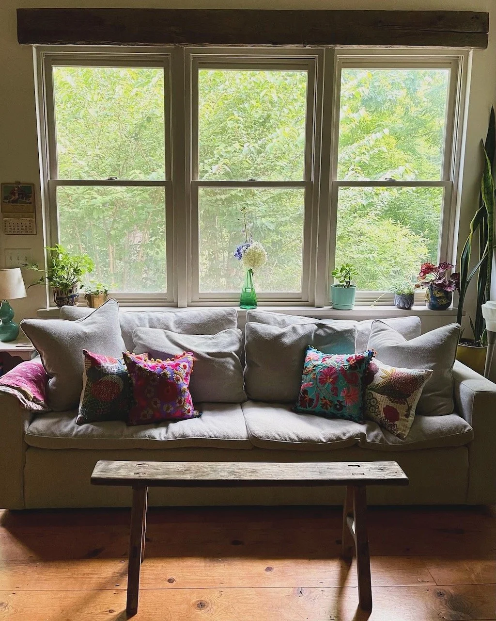 A cozy living room with a white sofa adorned with colorful patterned pillows, set in front of three large window panes showing lush green foliage outside. Floral decorations and houseplants sit on the window sill and beside the sofa, with a rustic wo