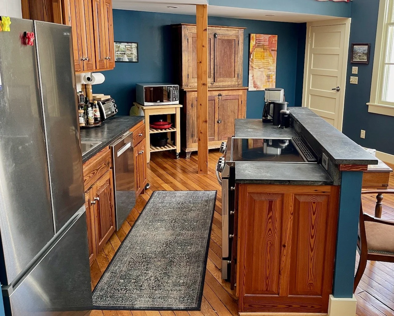 A kitchen featuring wooden cabinets, a stainless steel refrigerator, black countertops, and a wooden floor. There is a patterned rug on the floor, and various kitchen appliances on the counter, with a blue wall behind.