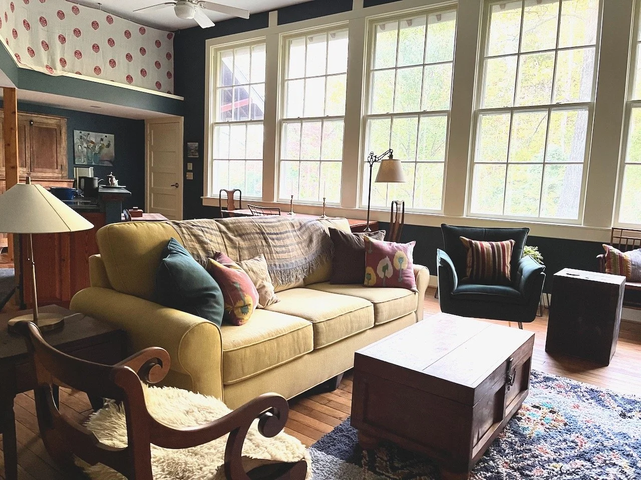 Living room with yellow sofa, black armchair, wooden coffee table, and large windows showing trees outside.