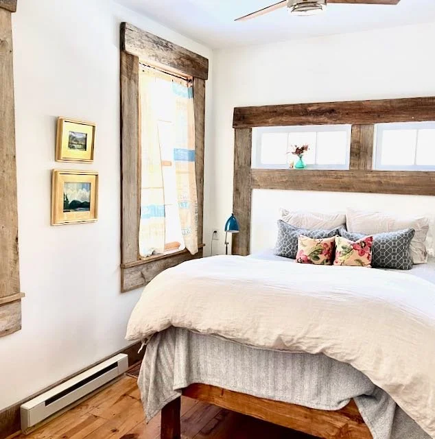 Cozy bedroom with white walls, wooden accents around a window and bed headboard, a white bed with multiple decorative pillows, a small blue bedside lamp, a vase with flowers on the headboard, and two small framed landscape pictures on the wall.