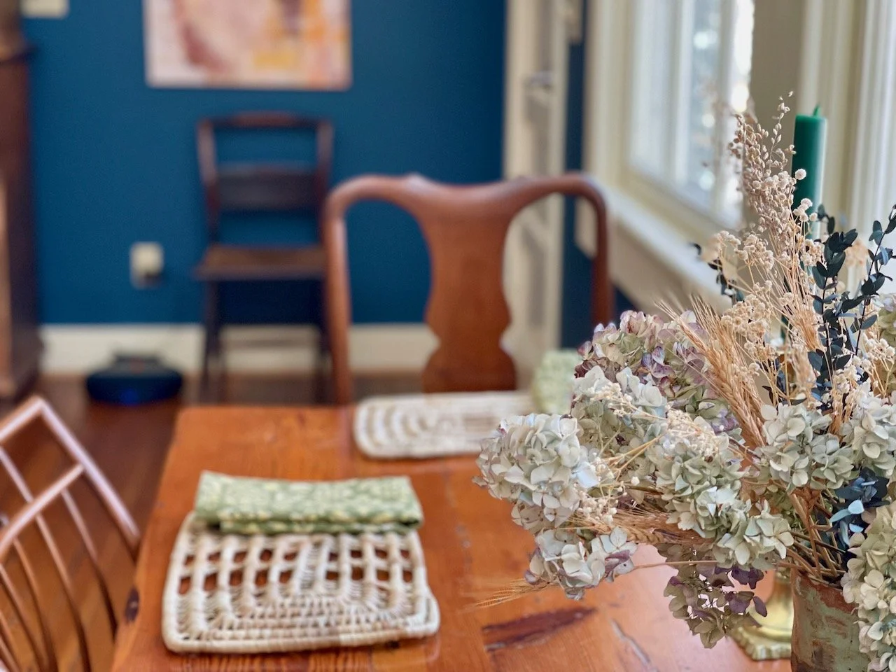 A close-up of a floral arrangement with dried and fresh flowers on a wooden dining table, with placemats and napkins. In the background, a rocking chair and a wooden chair are visible, with a blue wall and window letting in natural light.