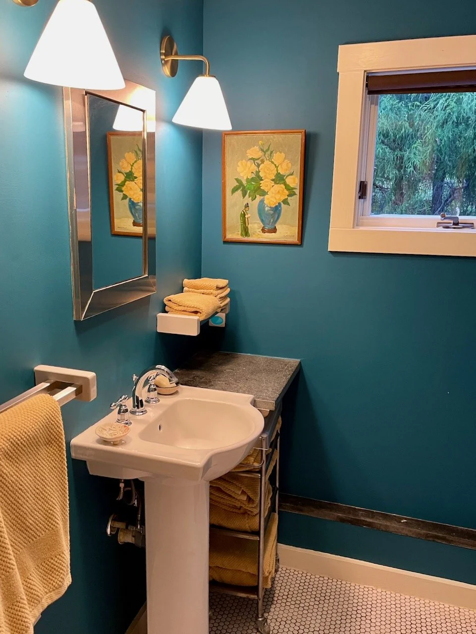 A small bathroom with blue walls, a white pedestal sink, a yellow towel hanging on a towel bar, a small mirror, wall-mounted light fixtures, a window showing greenery outside, a framed floral painting, and a metal shelf with yellow towels underneath 