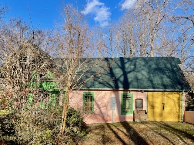 A small house with a green roof, pink and yellow walls, green window frames, and a green door, surrounded by leafless trees and bushes, under a clear blue sky.