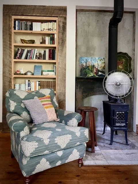 Living room corner with patterned armchair, bookshelf, small wooden stool, painting, and vintage fan, next to a stove and textured wall.
