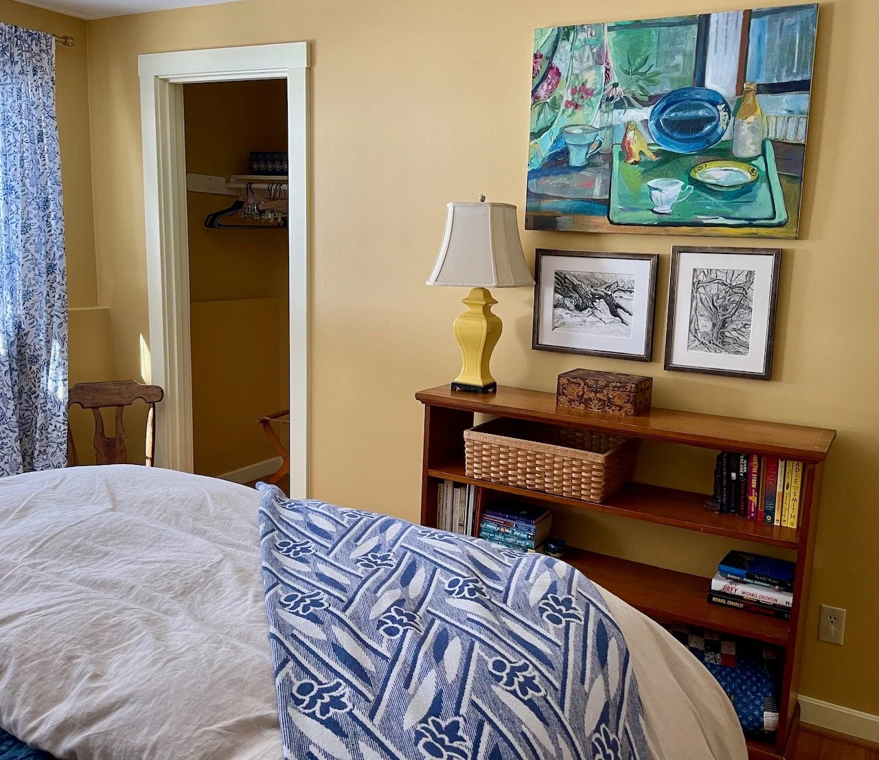 A cozy bedroom corner with a bed covered in a white and blue patterned blanket, a wooden bookshelf with books and a wicker basket, a yellow table lamp, and wall art including a colorful still life painting and two black-and-white sketches. A window w