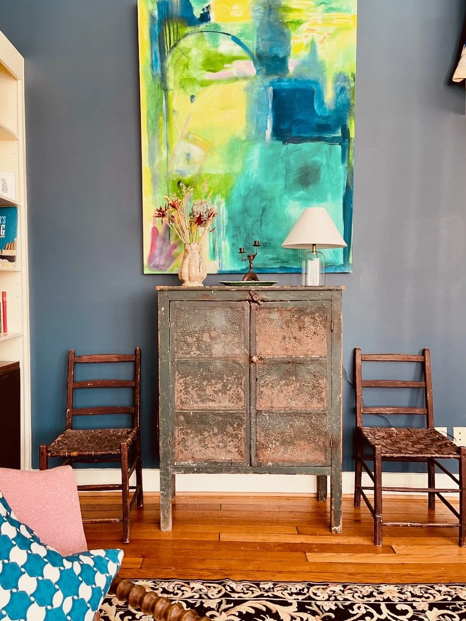 A rustic cabinet with a distressed finish is placed against a blue wall, decorated with a vase of flowers, a lamp, and a candelabra on top. Two wooden chairs with woven seats are positioned on either side of the cabinet. A large, abstract painting with bright colors hangs above it. The scene includes a patterned rug and part of a floral armchair in the foreground.