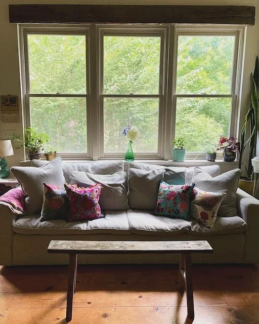 Living room with a cream sofa decorated with colorful pillows, positioned in front of three large windows with a view of green trees outside. A small wooden table is in front of the sofa, and potted plants sit on the window ledge, with a vase of flow