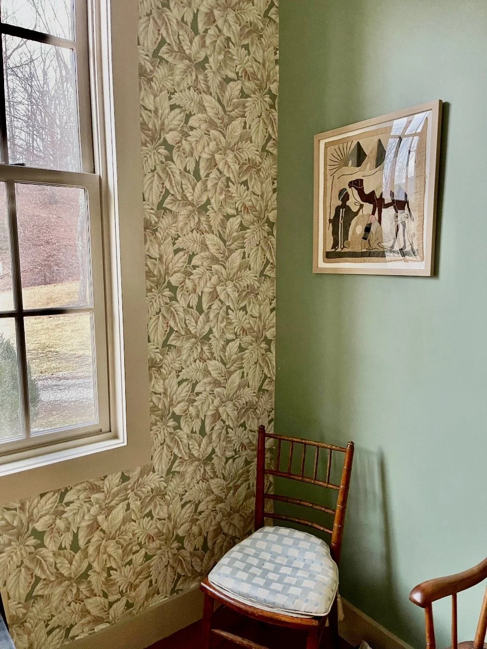 Corner of a room with a window, patterned wallpaper, a green wall, a framed artwork, and a wooden chair with a cushioned seat.