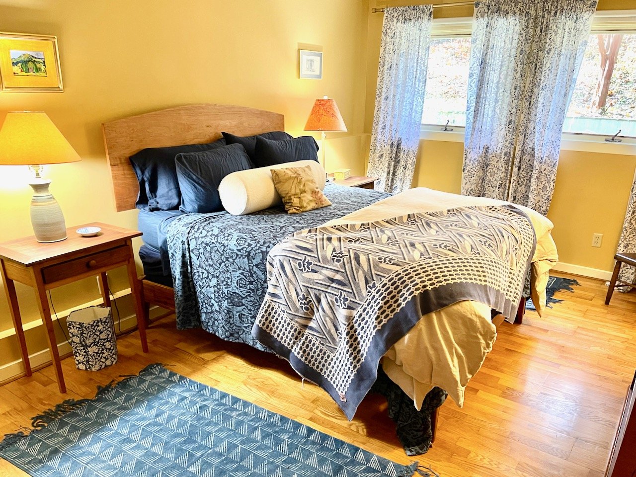 A neatly made bed with a wooden headboard, black pillows, a cylindrical bolster pillow, and decorative blankets in a cozy bedroom. There are two side tables with lamps, a patterned rug on the wooden floor, and windows with floral curtains letting in natural light.