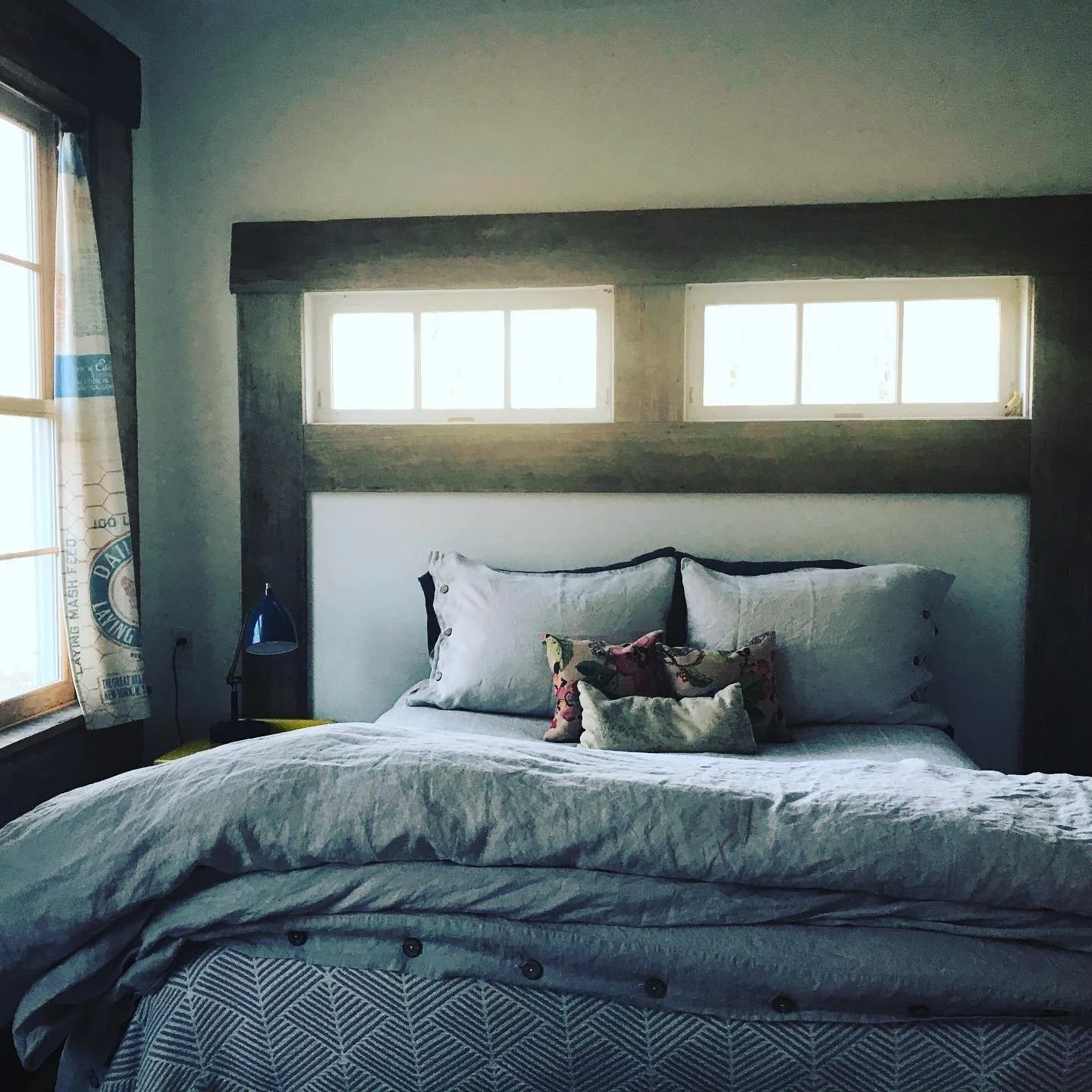 A cozy bedroom with a large bed, white bedding, multiple decorative pillows, a small bedside table, a black desk lamp, and two rectangular windows above the bed's headboard, with a curtain on the left window.