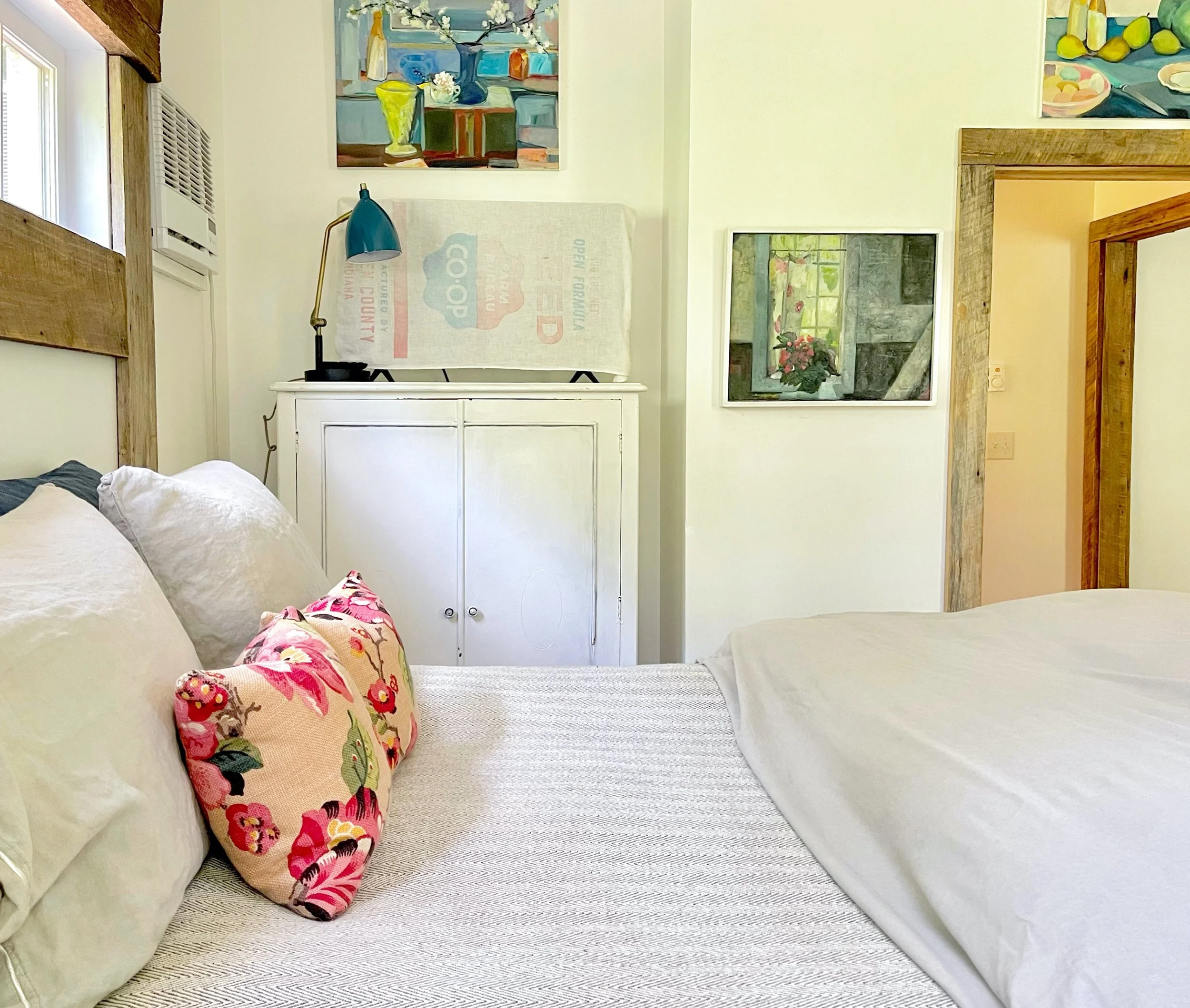 A cozy bedroom with a bed in the foreground, decorated with a pink floral pillow, white and beige pillows, a white textured blanket, and white sheets. In the background, there are colorful abstract paintings on the walls, a blue desk lamp on a white 