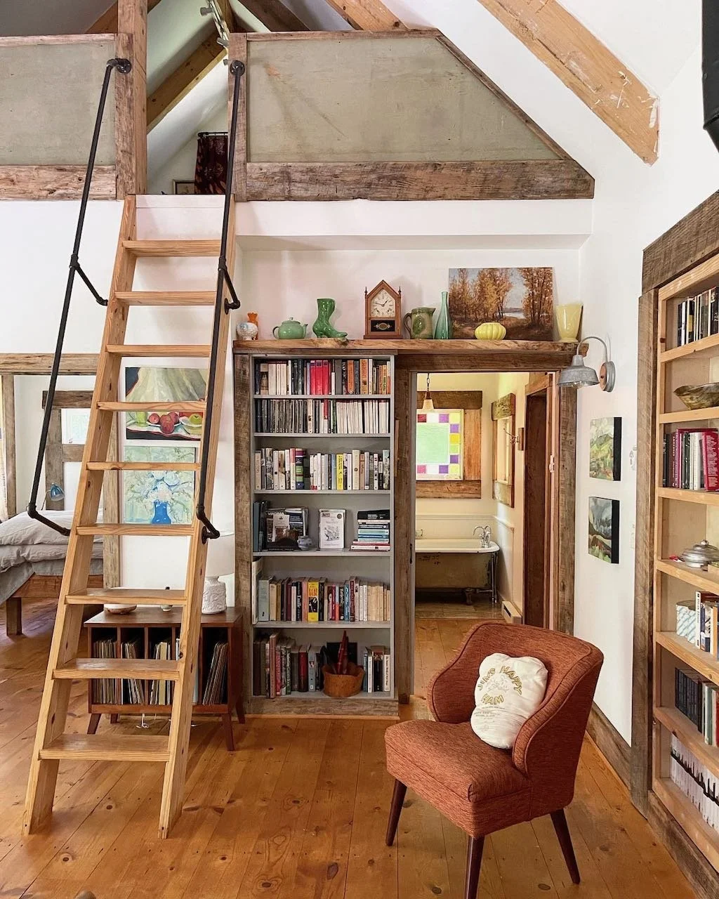 Living room with wooden beams, a wooden ladder leading to a loft, a bookshelf filled with books, a cushioned armchair, and decorative items on shelves.