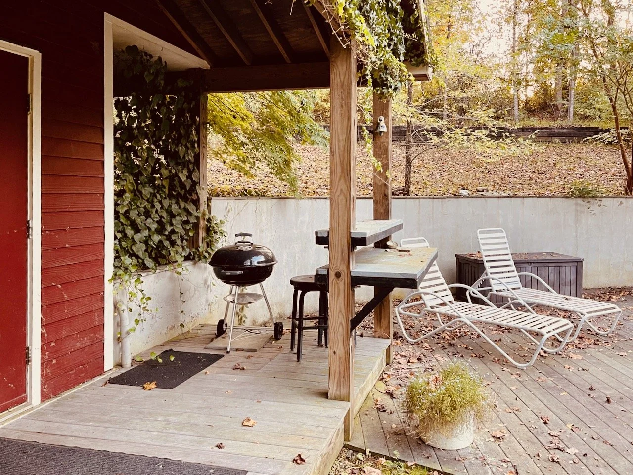 Backyard patio with two white lounge chairs, a black charcoal grill, a small table, a storage box, and a potted plant on weathered wooden decking, surrounded by trees with autumn leaves.