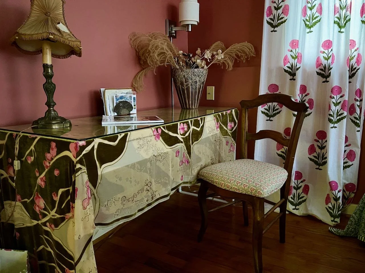 A vintage wooden chair with a patterned cushioned seat next to a decorated dresser with a floral tablecloth, a glass vase with dried pampas grass and white flowers, a table lamp with a fringed shade, and a sliding door with floral curtains in a pink-